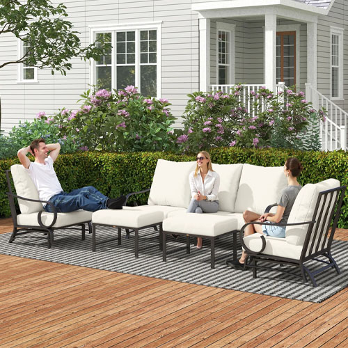 Outdoor patio seating set with metal frames and white cushions, featuring a loveseat, two armchairs, a coffee table, and an ottoman on a deck with floral and leafy surroundings.