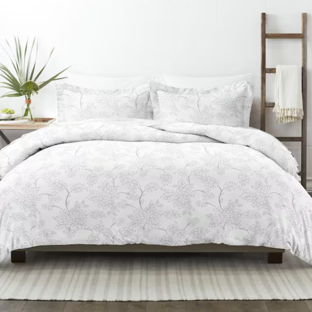 A white floral-patterned bedding set on a bed, accompanied by two matching pillowcases. The setting includes a side table with a vase and a ladder with a draped blanket.