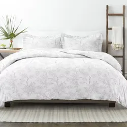 A white floral-patterned bedding set on a bed, accompanied by two matching pillowcases. The setting includes a side table with a vase and a ladder with a draped blanket.