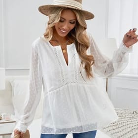 A white V-neck blouse with long sleeves, featuring a dotted pattern and lace trim details. The top is paired with a straw hat and layered necklaces, creating a light, stylish look.