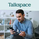 A man sits in a cozy room holding a smartphone, with the logo \“Talkspace\“ displayed above, suggesting a focus on digital communication or mental health services.