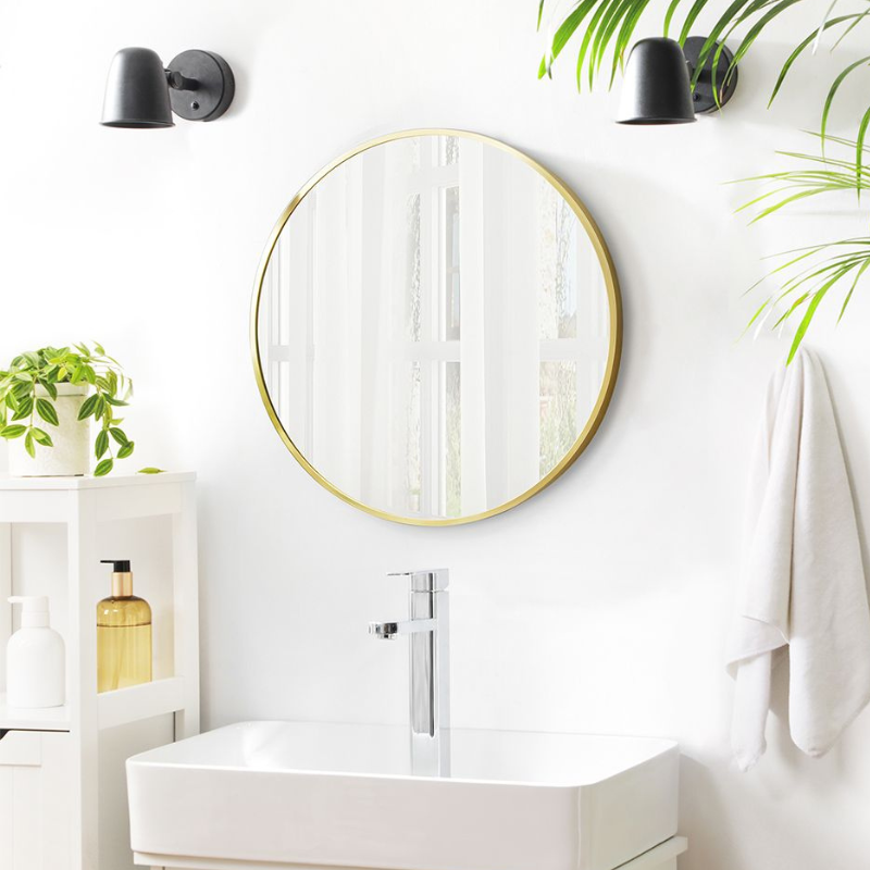 A bathroom setup featuring a round mirror with a gold frame, modern faucet, and wall sconces. It includes a small shelf with plants and bottles, set against a bright, white background.