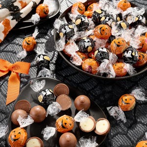 Assorted Lindt Lindor Halloween truffles are displayed in decorative wrappers featuring pumpkin and ghost designs, presented in black bowls on a textured tablecloth.