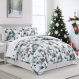 A cozy bedroom features a winter-themed comforter set with tree and house patterns. The room is decorated for Christmas with a tree, stockings, and wrapped gifts, complemented by a snowy forest painting and neutral decor.