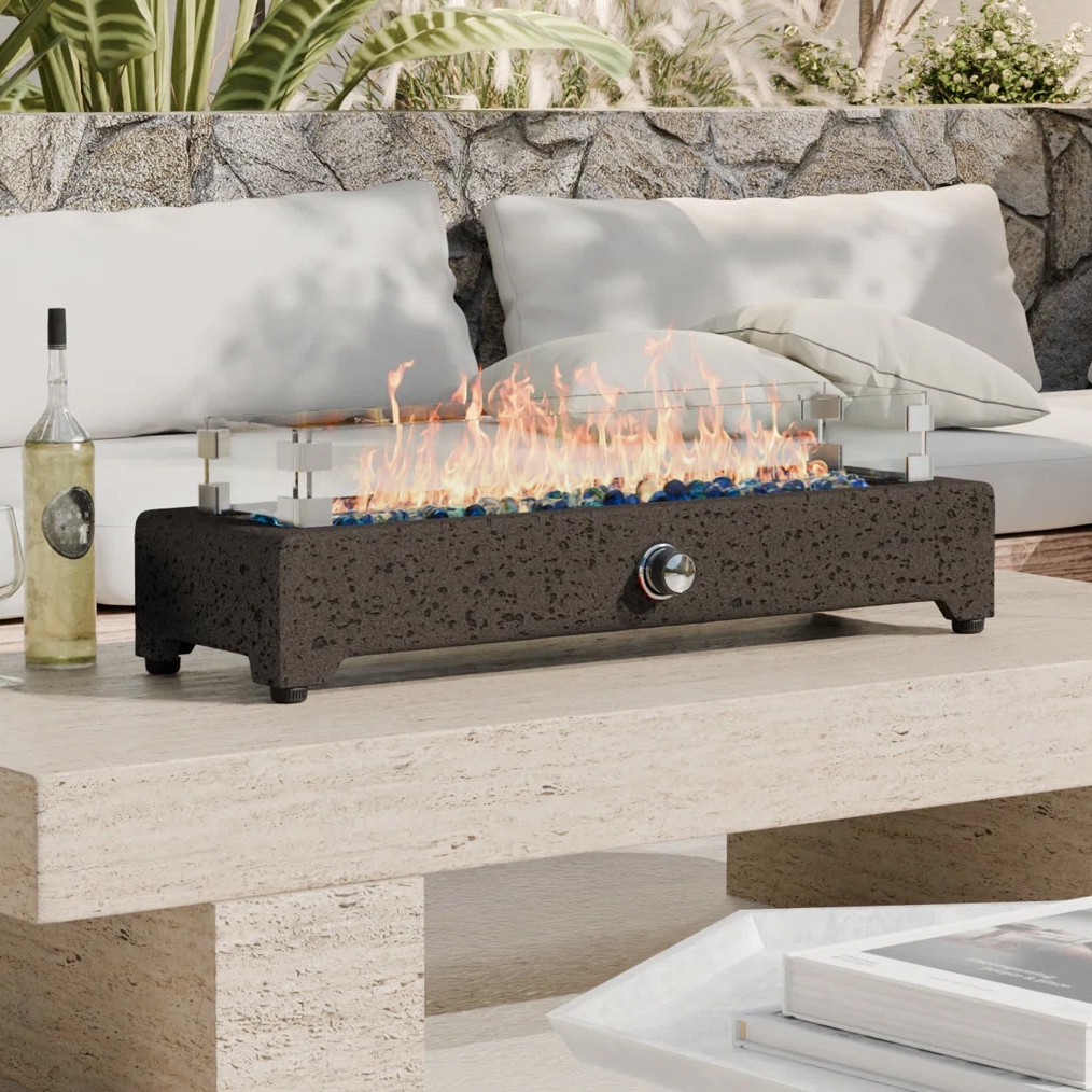 A tabletop fire pit with a glass wind guard sits on a stone table next to an outdoor sofa. A wine bottle, glass, and a magazine are also on the table.