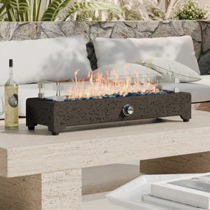 A tabletop fire pit with a glass wind guard sits on a stone table next to an outdoor sofa. A wine bottle, glass, and a magazine are also on the table.