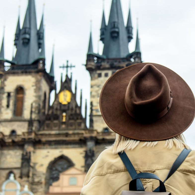 A person wearing a brown wide-brimmed hat and a beige jacket stands in front of a historic church with tall spires, featuring a light gray backpack.