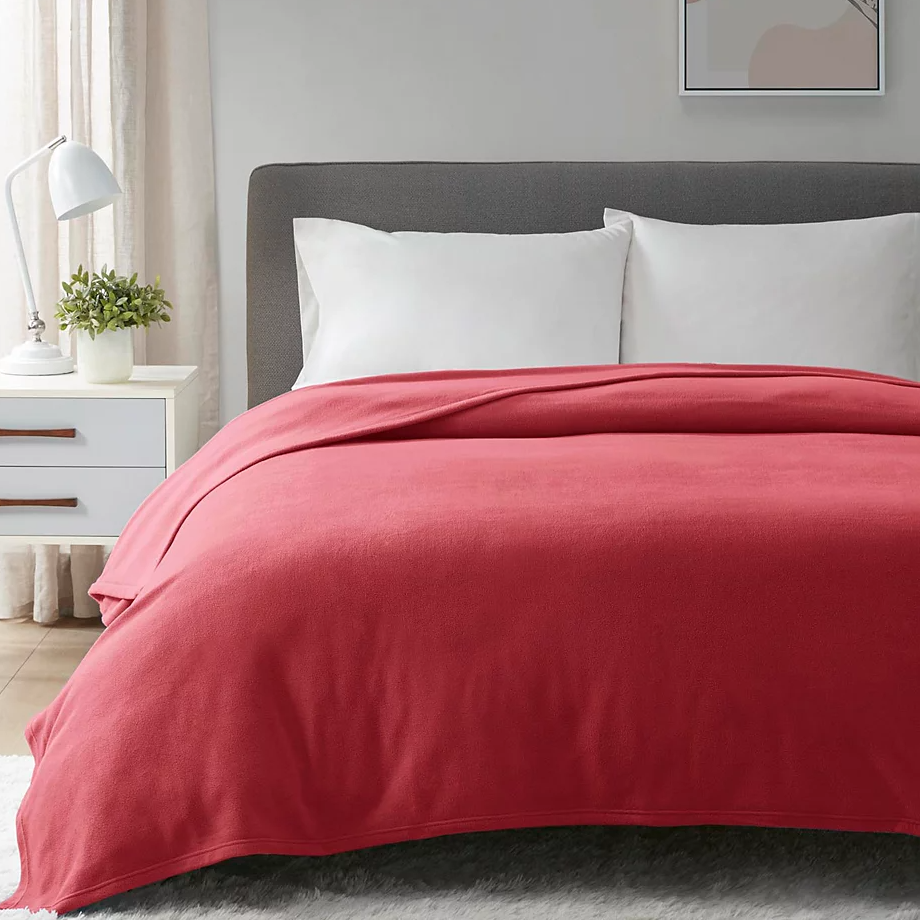 A neatly made bed with a coral-colored comforter, white pillows, and a gray headboard is shown. Nearby, there's a white nightstand with a lamp and a small plant.