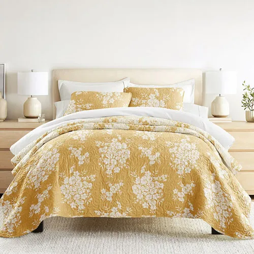 A 3-piece all-season queen quilt set featuring a mustard and white floral pattern, including two matching pillow shams. It's displayed in a minimalistic bedroom setup with beige lamps, a light wood nightstand, and a soft gray rug.