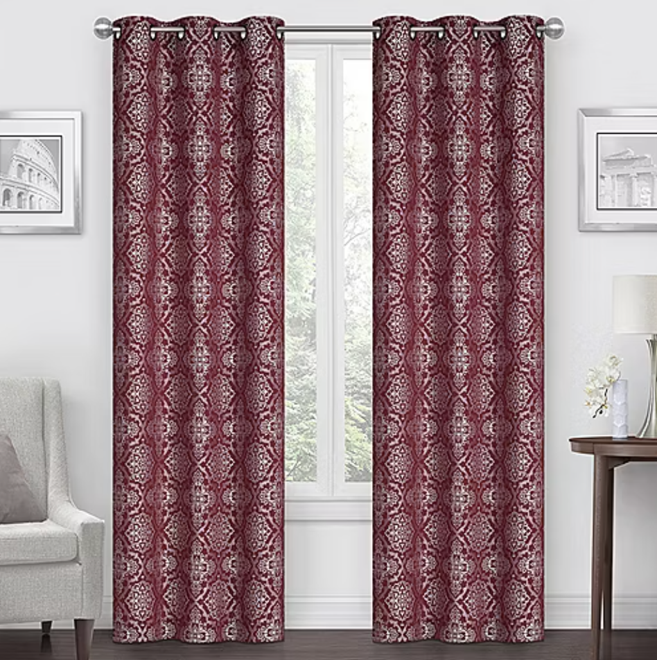 Two patterned burgundy curtains with silver grommets hang in a living room setting, framed by a window. They complement a light-colored chair, wall art, and a side table with a lamp.