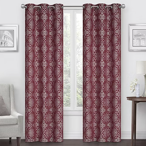 Two patterned burgundy curtains with silver grommets hang in a living room setting, framed by a window. They complement a light-colored chair, wall art, and a side table with a lamp.
