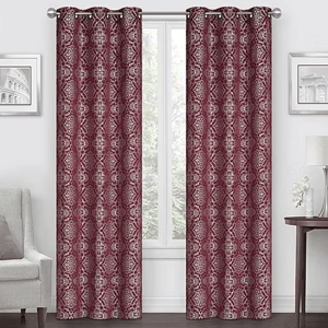 Two patterned burgundy curtains with silver grommets hang in a living room setting, framed by a window. They complement a light-colored chair, wall art, and a side table with a lamp.