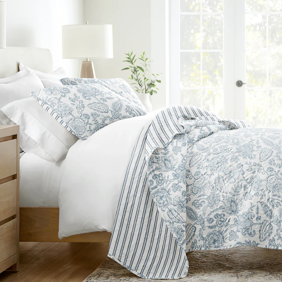 A bed is adorned with white and blue floral-patterned bedding, featuring a duvet and pillowcases with coordinating stripes. A wooden nightstand with a lamp and books is beside the bed, and a large window allows natural light to fill the room.