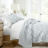 A bed is adorned with white and blue floral-patterned bedding, featuring a duvet and pillowcases with coordinating stripes. A wooden nightstand with a lamp and books is beside the bed, and a large window allows natural light to fill the room.