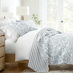 A bed is adorned with white and blue floral-patterned bedding, featuring a duvet and pillowcases with coordinating stripes. A wooden nightstand with a lamp and books is beside the bed, and a large window allows natural light to fill the room.