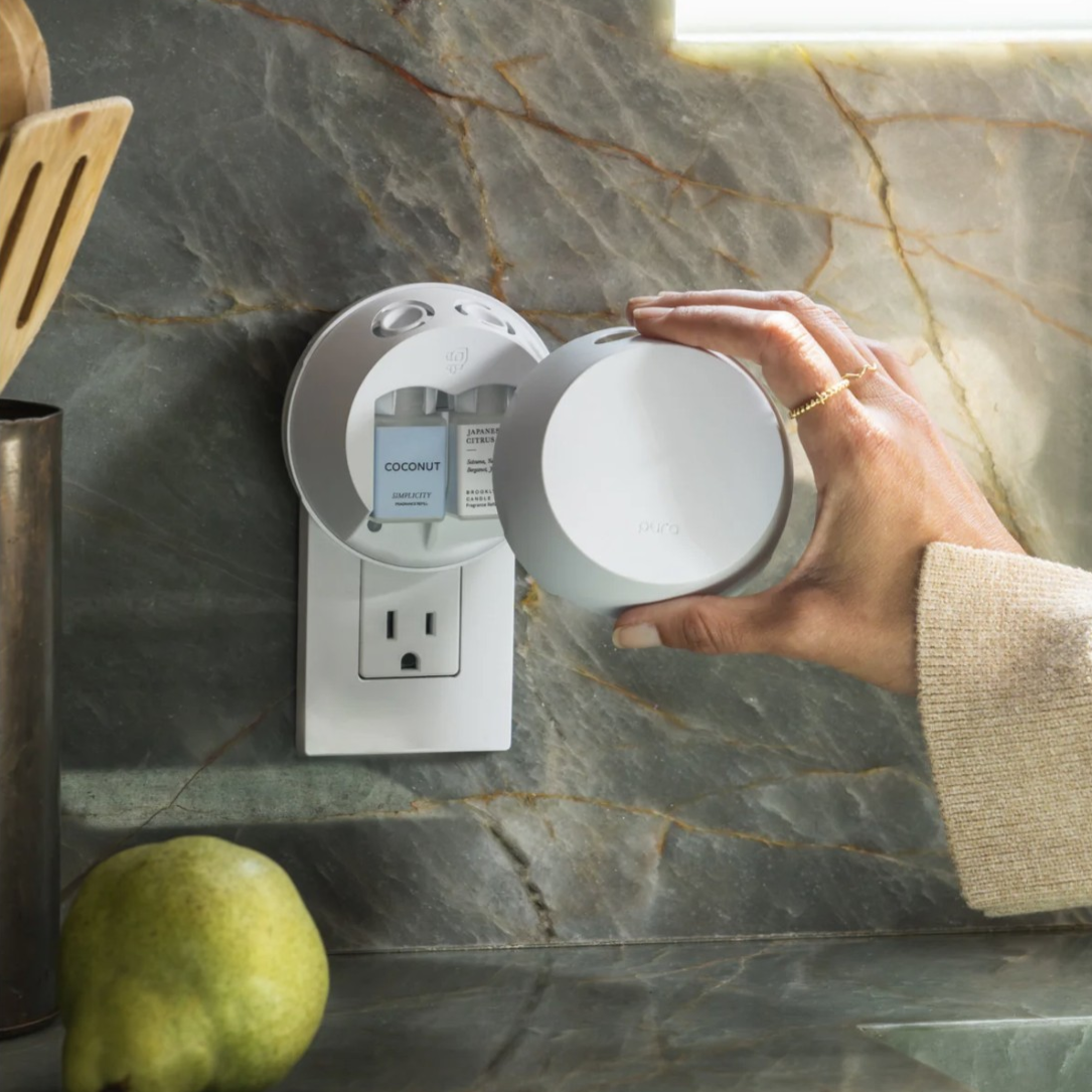 A person is placing a round Pura smart home fragrance diffuser into a wall outlet with a pear nearby on a marble countertop.