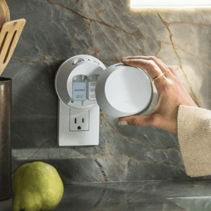 A person is placing a round Pura smart home fragrance diffuser into a wall outlet with a pear nearby on a marble countertop.