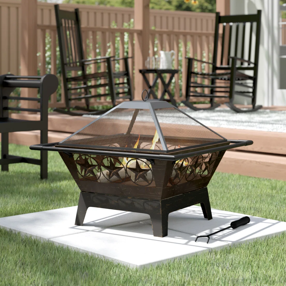 Outdoor fire pit with star designs, set on a grass lawn, surrounded by black rocking chairs and a wooden deck with flower background.