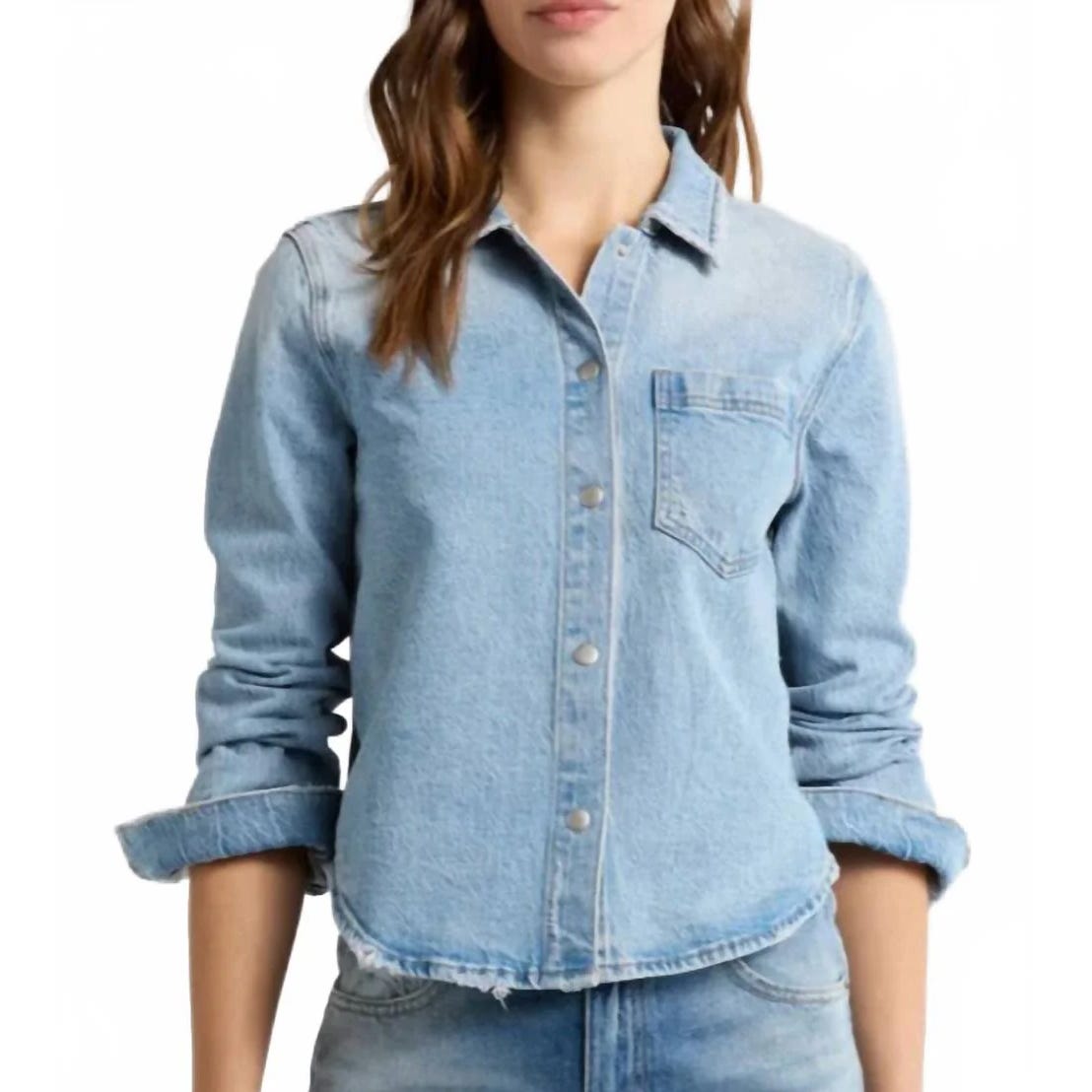 A woman is wearing a light blue denim button-up shirt with a single chest pocket and rolled-up sleeves.