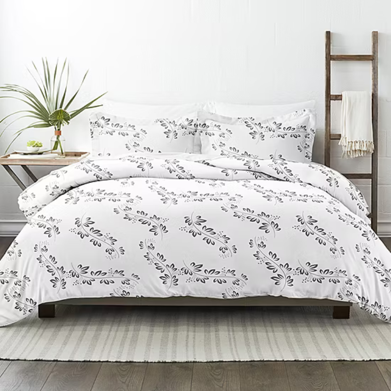 White bedding set with a black floral pattern, featuring a duvet cover and pillowcases. It is displayed on a bed with a wooden frame, accompanied by a side table with green plants and a decorative ladder with a cream-colored blanket.