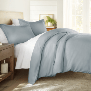 A light blue 300-thread count cotton duvet cover set on a bed, accompanied by matching pillowcases. The setting includes a wooden side table with decor and a bright window in the background.