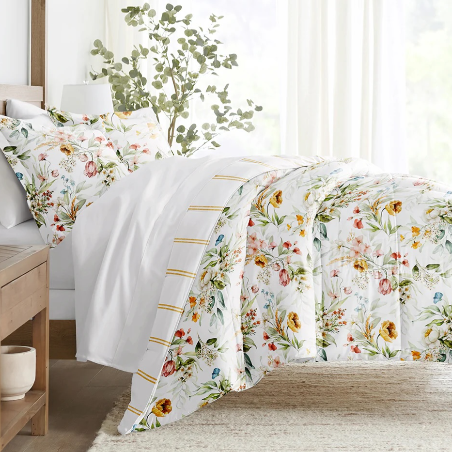 A neatly made bed with a floral-patterned duvet cover and matching pillowcases sits in a bright room. A small wooden side table with a ceramic pot is nearby, with greenery visible through sheer curtains.