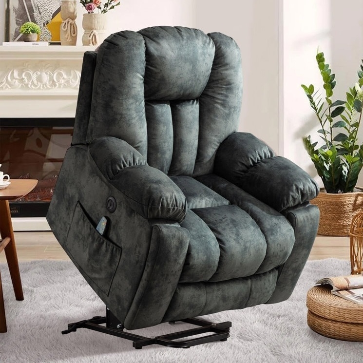A plush, dark gray reclining lift chair with a side pocket contains a remote control, placed in a cozy living room setting with plants and a fireplace.