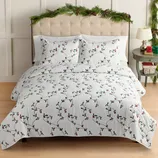 A bed is covered with white bedding featuring a design of green vines and red berries. The headboard is decorated with festive greenery, and the nightstand holds a lamp and a small wrapped gift.