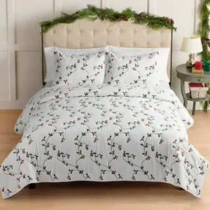 A bed is covered with white bedding featuring a design of green vines and red berries. The headboard is decorated with festive greenery, and the nightstand holds a lamp and a small wrapped gift.