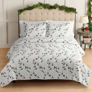 A bed is covered with white bedding featuring a design of green vines and red berries. The headboard is decorated with festive greenery, and the nightstand holds a lamp and a small wrapped gift.