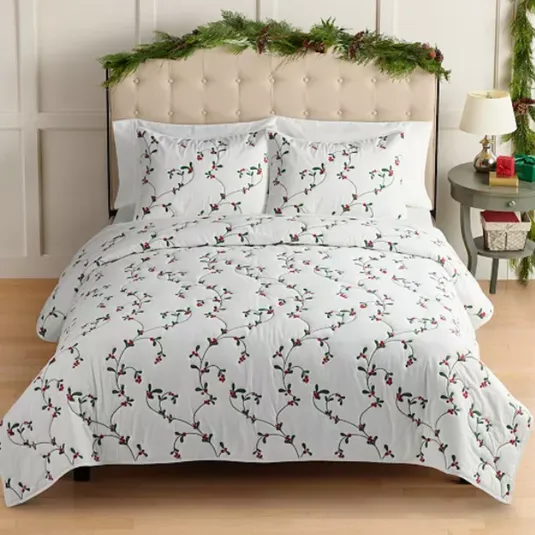 A bed is covered with white bedding featuring a design of green vines and red berries. The headboard is decorated with festive greenery, and the nightstand holds a lamp and a small wrapped gift.