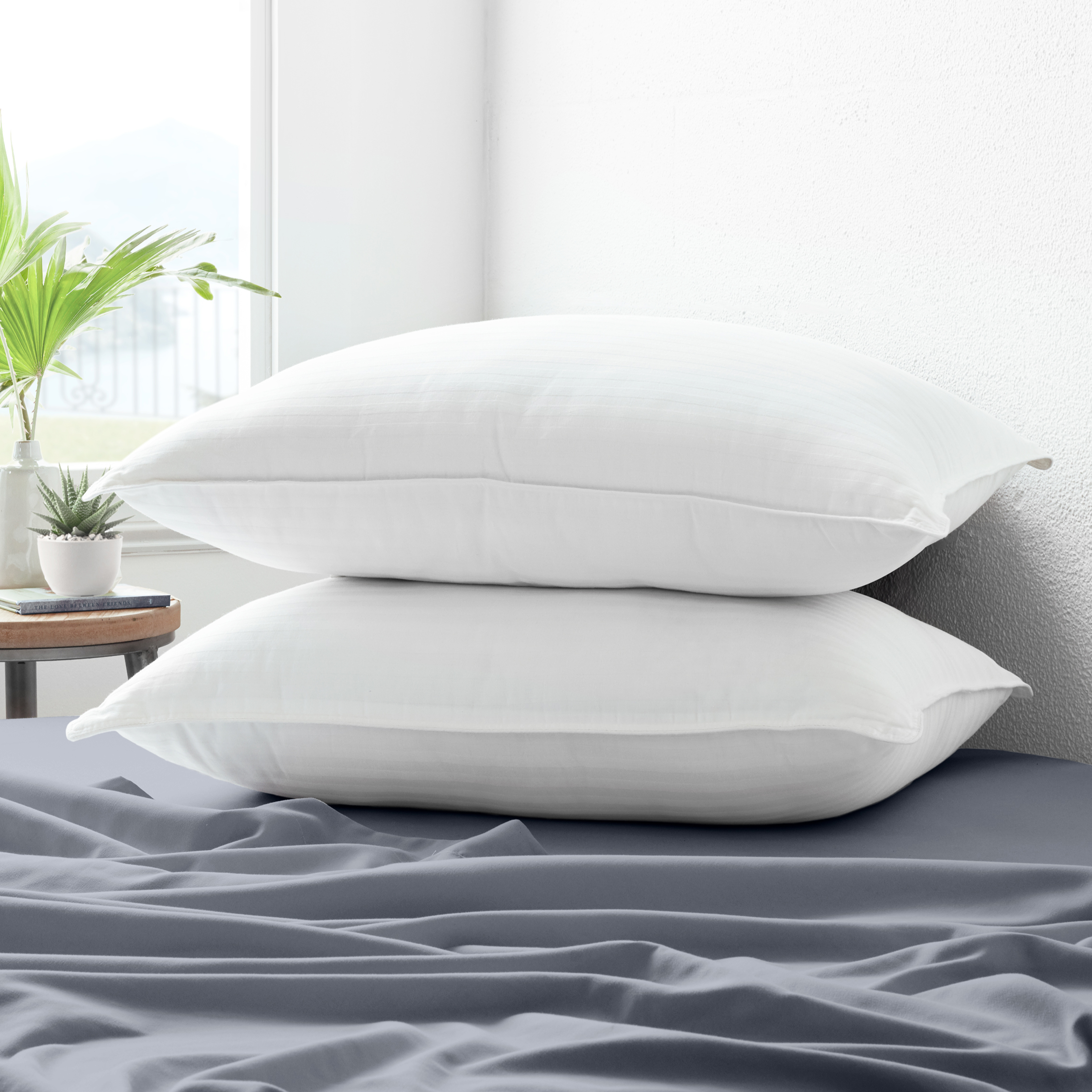 Two white pillows on a grey bedsheet, with a small plant and decorative items on a bedside table in the background.