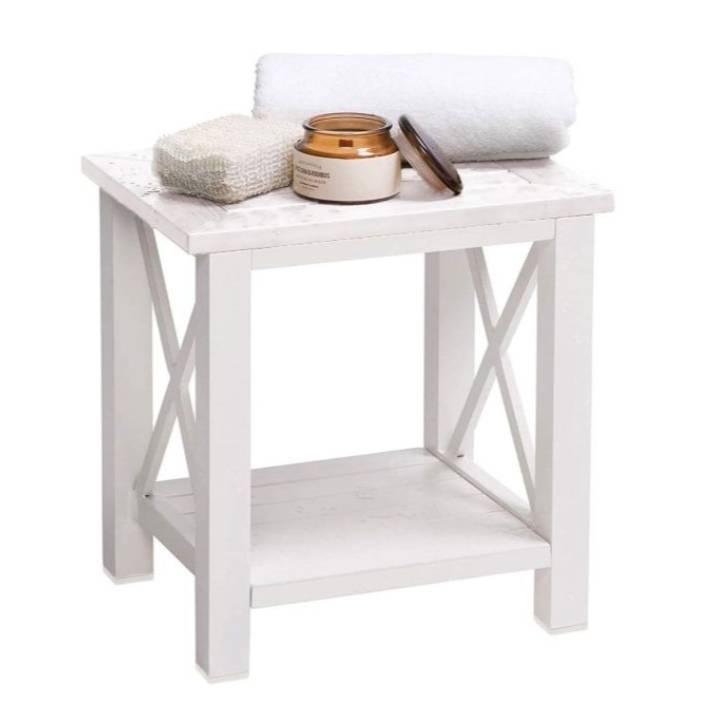 A white wooden bench with a slatted top and an X-frame design on the sides, featuring a lower shelf, a towel, and toiletry items on top.