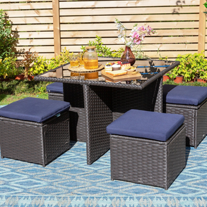 Outdoor rattan patio set with four cushioned cube stools, a glass-top table, and decorative items like a floral vase, a tray of cupcakes, and a jug with glasses, on a blue-patterned rug in a garden setting.