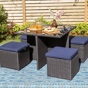 Outdoor rattan patio set with four cushioned cube stools, a glass-top table, and decorative items like a floral vase, a tray of cupcakes, and a jug with glasses, on a blue-patterned rug in a garden setting.