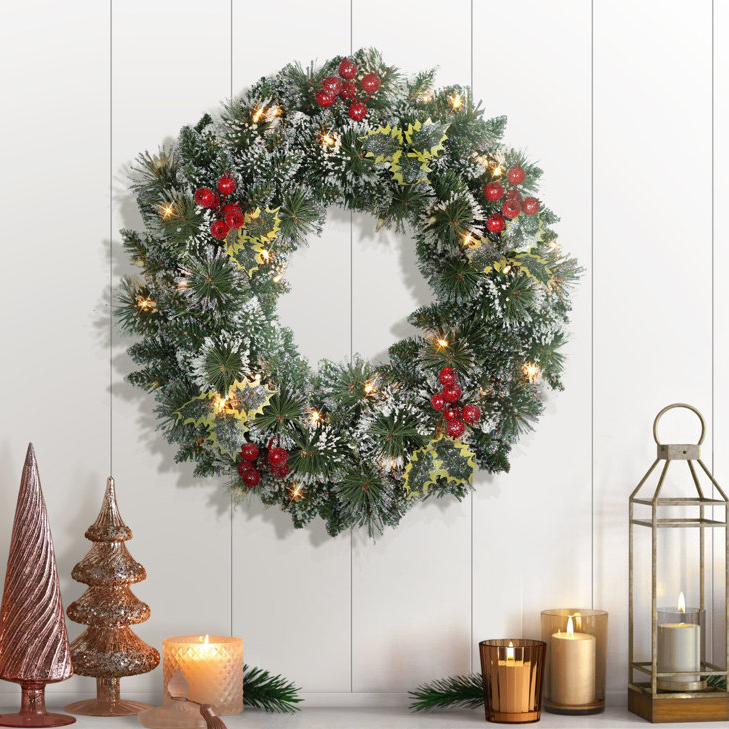 A festive wreath adorned with red berries, holly leaves, and warm white lights hangs on a wall, accompanied by candles and a lantern on the side.