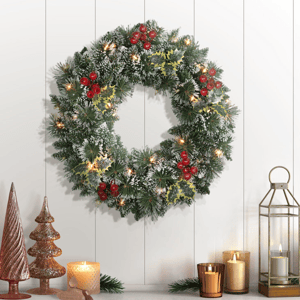 A festive wreath adorned with red berries, holly leaves, and warm white lights hangs on a wall, accompanied by candles and a lantern on the side.