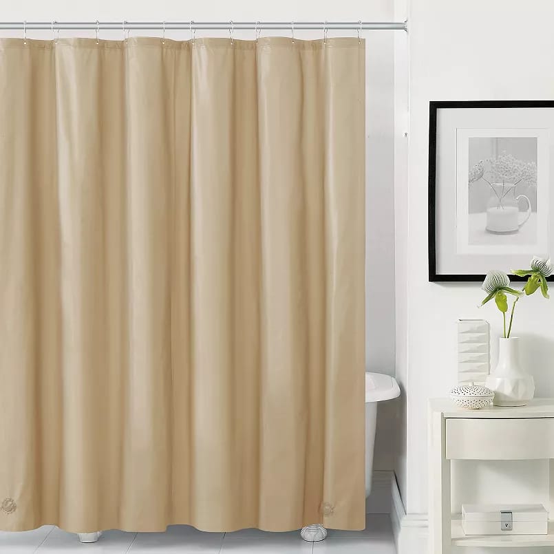 A plain beige shower curtain liner is shown, hanging from a shower rod, with a white shelf, a plant, and a framed picture in the background.