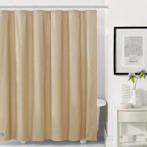 A plain beige shower curtain liner is shown, hanging from a shower rod, with a white shelf, a plant, and a framed picture in the background.