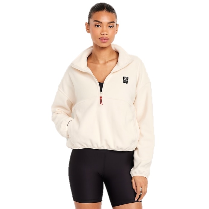 A woman is wearing a beige fleece quarter-zip pullover with a logo patch on the chest and black cycling shorts, standing against a white background.