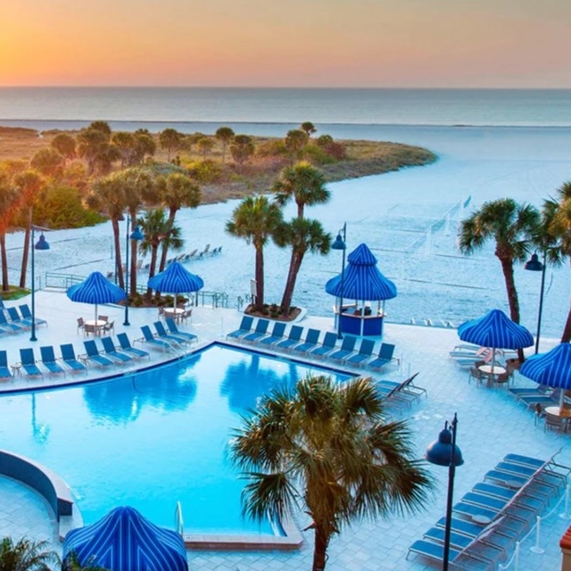 A beach resort pool area with palm trees, blue lounge chairs, and blue-striped umbrellas, overlooking a pristine beach and ocean at sunset.