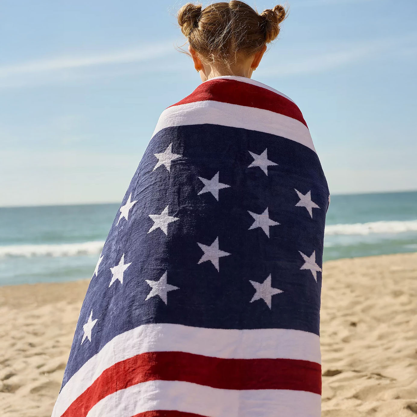 A person on the beach is wrapped in an American flag-themed towel, featuring red, white, and blue stripes with white stars.