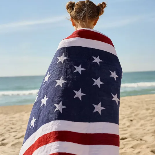 A person on the beach is wrapped in an American flag-themed towel, featuring red, white, and blue stripes with white stars.