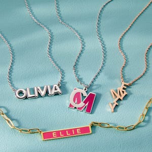 Assorted custom necklaces with personalized names \“OLIVIA,\“ \“ELLIE,\“ and initials \“A\“ and \“AAA\“ in various fonts and colors, displayed on a teal background.