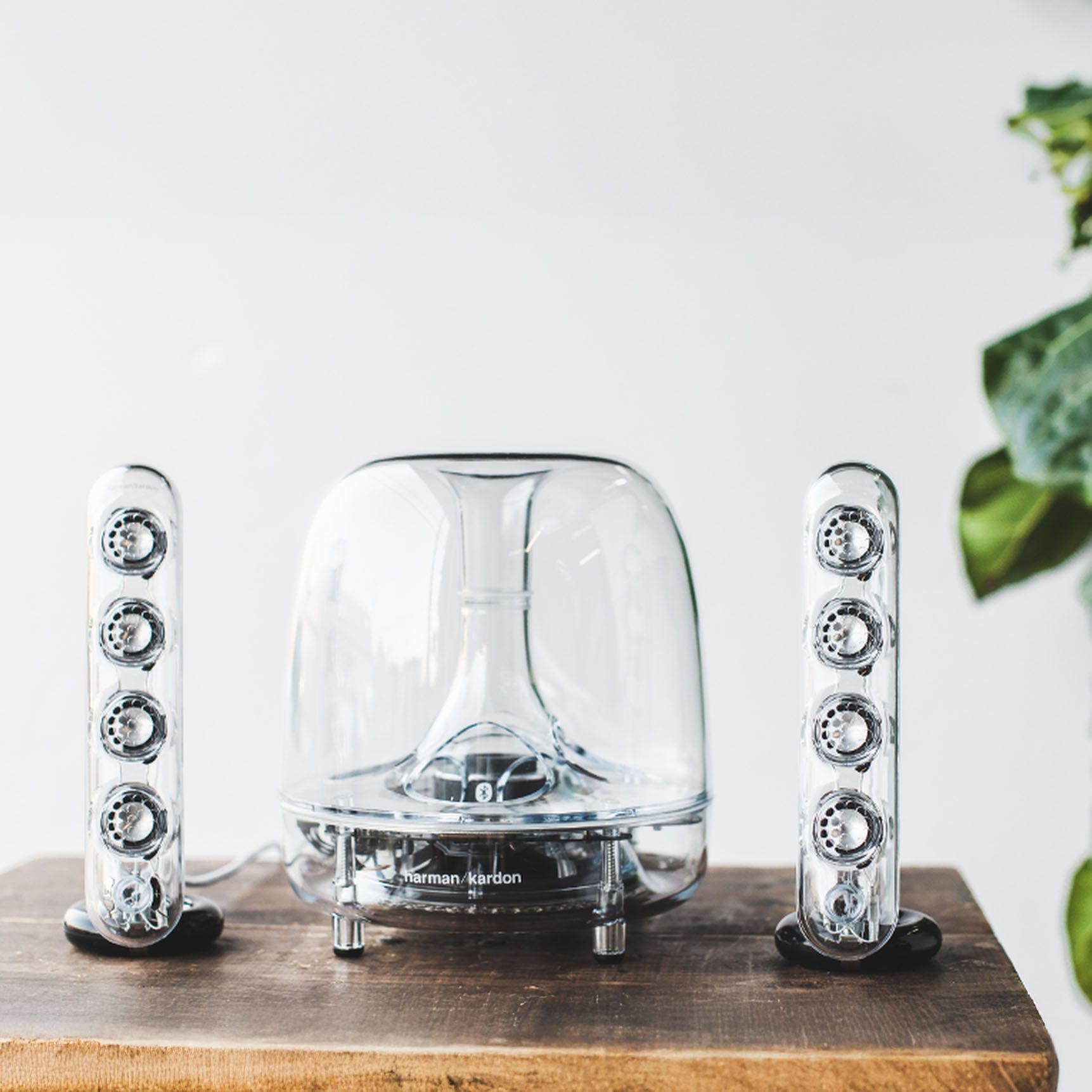 Harman Kardon transparent speaker set with a dome-shaped subwoofer and two vertical satellite speakers on a wooden surface.