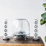 Harman Kardon transparent speaker set with a dome-shaped subwoofer and two vertical satellite speakers on a wooden surface.
