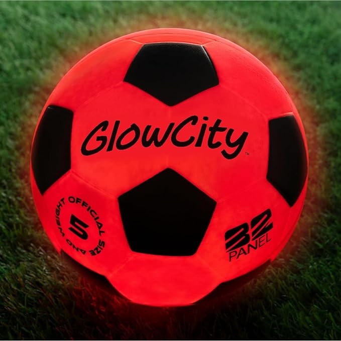 A red glow-in-the-dark soccer ball with black panels, featuring \“GlowCity\“ branding and labeled as an official size 5 with 32 panels, set on grass.