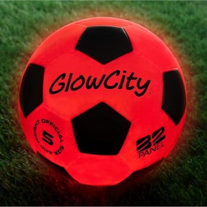A red glow-in-the-dark soccer ball with black panels, featuring \“GlowCity\“ branding and labeled as an official size 5 with 32 panels, set on grass.