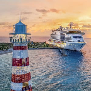 A large cruise ship is docked near a lighthouse with a red and white pattern, against a scenic sunset backdrop.
