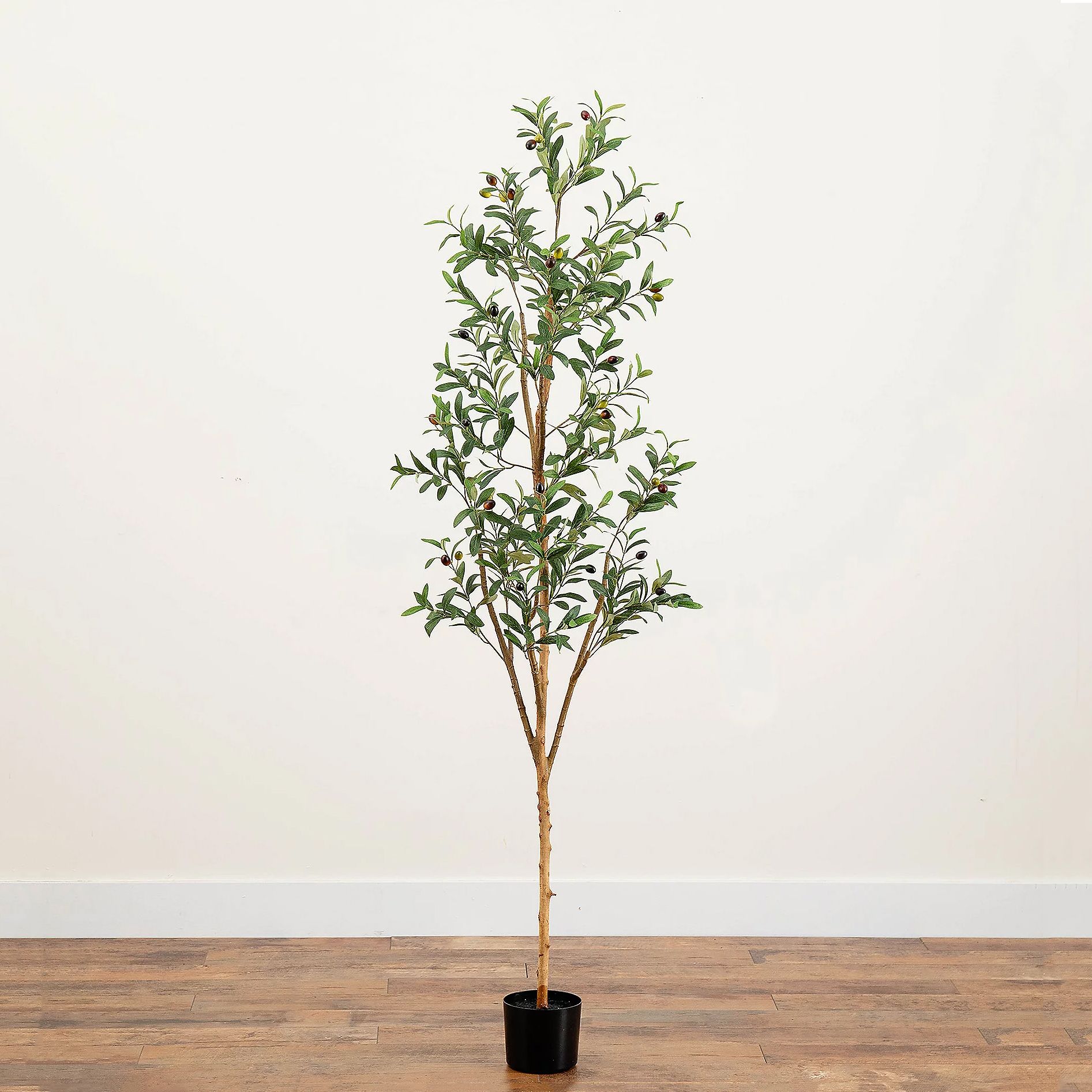 A 6-foot artificial olive tree with realistic green leaves and small olives, set in a black pot, stands on a wooden floor against a plain white wall.
